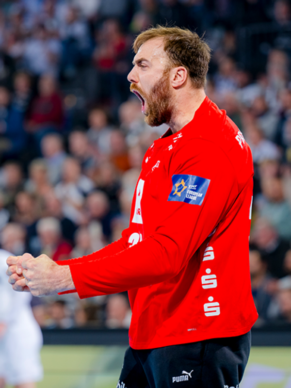 Der Torwart Andreas Wolff trägt ein langärmliges rotes Trikot, ballt die Fäuste und jubelt mit offenem Mund. Im Hintergrund ist die Tribüne mit Zuschauern