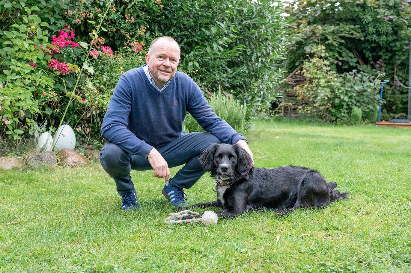 Ein Mann hockt mit seinem schwarzen Hund im Garten auf der Wiese. Im Hintergrund sind Büsche zu sehen.