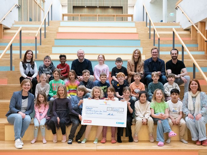 Gruppenfoto von Kindern und Erwachsenen auf einer großen Holztreppe in einem hellen Innenraum. In der Mitte halten mehrere Kinder einen großen Spendenscheck über 5.000 Euro mit REWE- und coop-Logo in die Kamera.