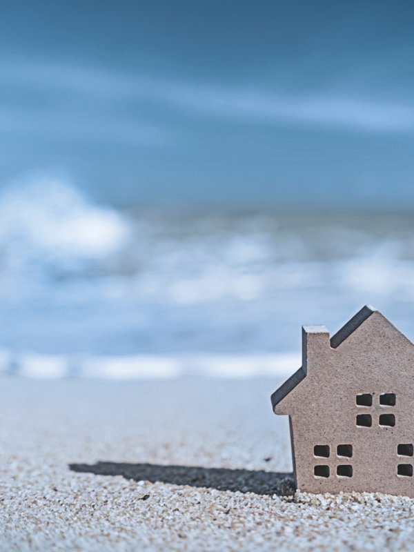 Rechts im Vordergrund ist ein kleines graues Haus, was im Sand am Strand steht. Im Hintergrund kommen die blauen Wasserwellen auf das Haus zu. 