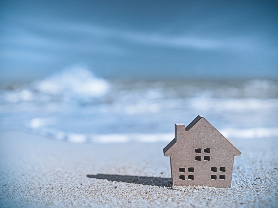 Rechts im Vordergrund ist ein kleines graues Haus, was im Sand am Strand steht. Im Hintergrund kommen die blauen Wasserwellen auf das Haus zu. 