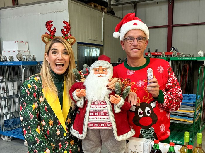 Frau mit Rentier-Haarreif und Mann mit Nikolausmütze posieren strahlend mit Deko-Weihnachtsmann bei Steffenhagen Großhandel, hinter einem der Weihnachtsmarkt-Stände.