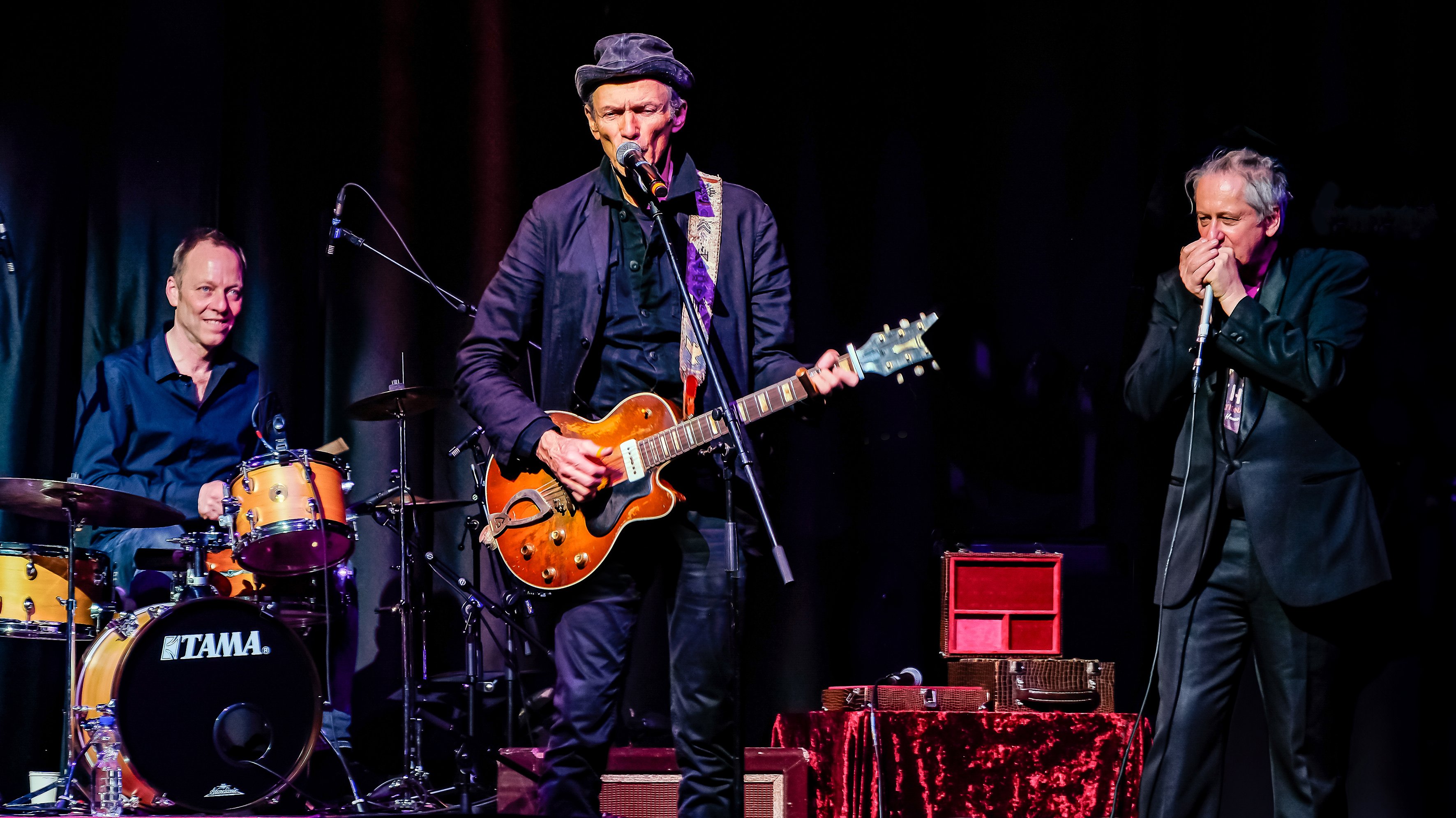 Dunkler Hintergrund, im Vordergrund ein zugleich singender und Gitarre spielender Mann mit Hut, links daneben ein Schlagzeuger, rechts daneben ein Mann der Mundharmonika spielt