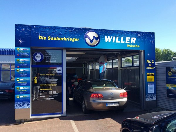 Ein Auto fährt gerade in die Waschanlage von Clean Car, die einen blauen Rahmen mit dem Schriftzug hat. Im Hintergrund ist blauer Himmel. 