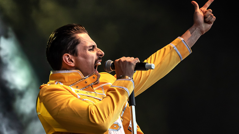 Freddy Mercury Double reißt in einem gelben Jacket den linken Arm in die Luft und hält in der rechten Hand ein Mikrofon an den Mund
