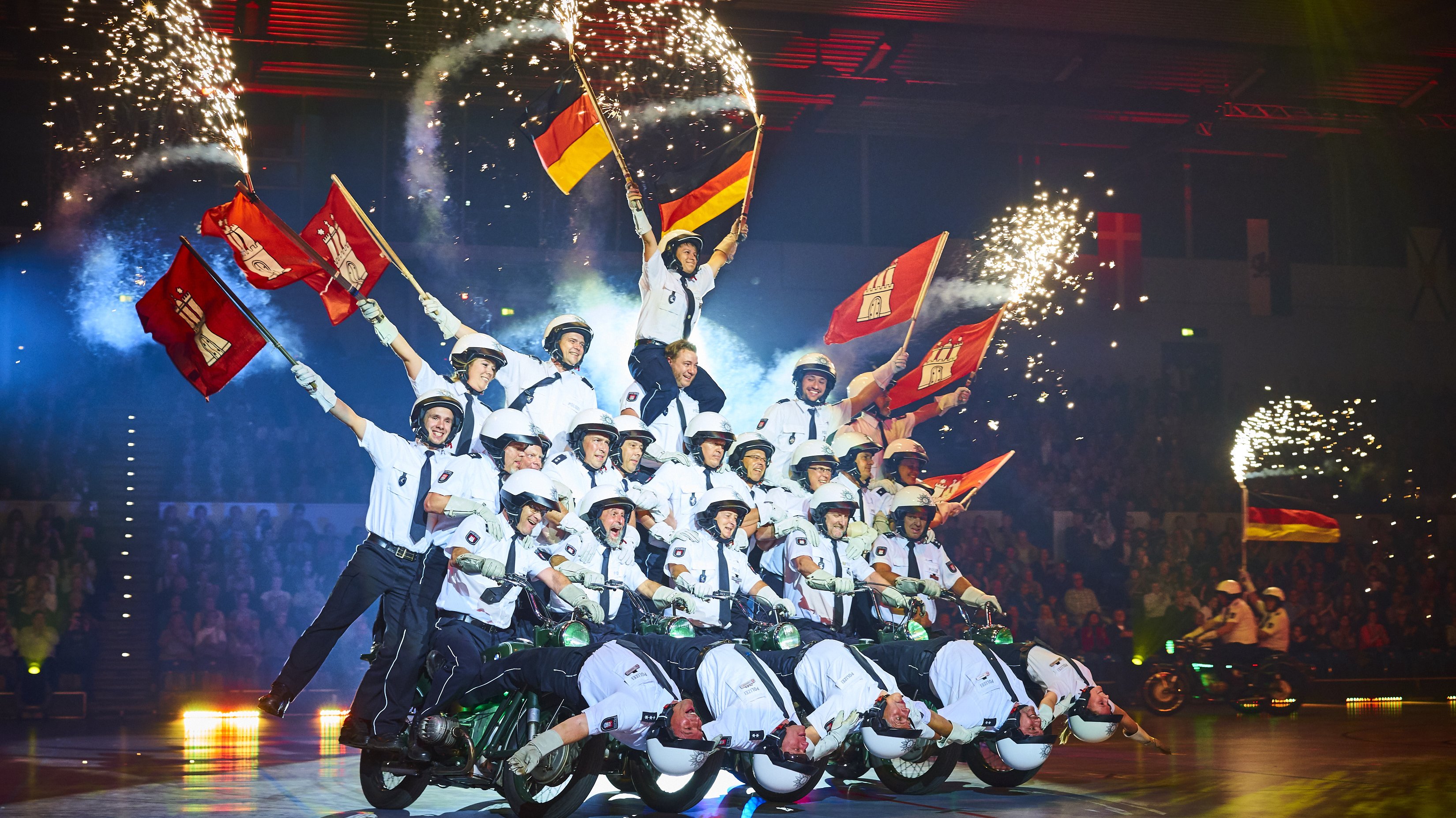 Pyramide mit Polizisten, die unterste Reihe sitzt auf Motorrädern, die Oberen schwenken Fahnen, Feuerwerk im Hintergrund