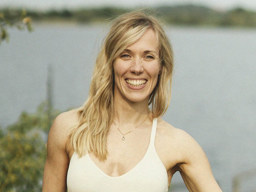 Eine blonde Frau mit breitem Lächeln und weißem Top. Im Hindergrund ist ein See