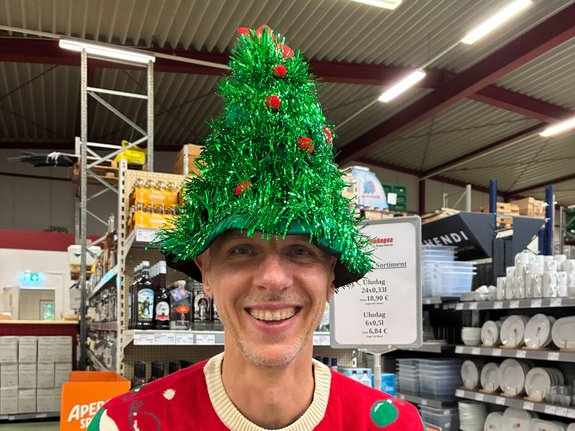 Mann mit glitzerndem Tannenbaum-Hut und rotem ‘MEOWY’-Weihnachtspullover lächelt zwischen Regalen im Großhandel Steffenhagen.