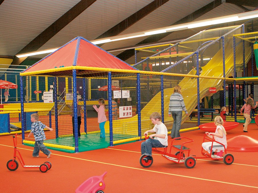 In dem Indoorspiel sind verschiedenen Spielgeräte, wie Fahrräder oder eine Rutsche zu sehen. Im Vordergrund fahren gerade drei Kinder mit dem Fahrrad. 