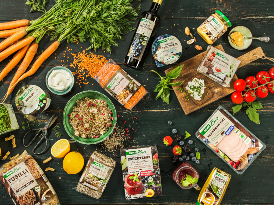 Auf einer Holzplatte liegen viele gesunde REWE-Bio Prokukte. Von Obst und Gemüse über Nudeln, Linsen, Wurst und Hüttenkäse ist alles dabei