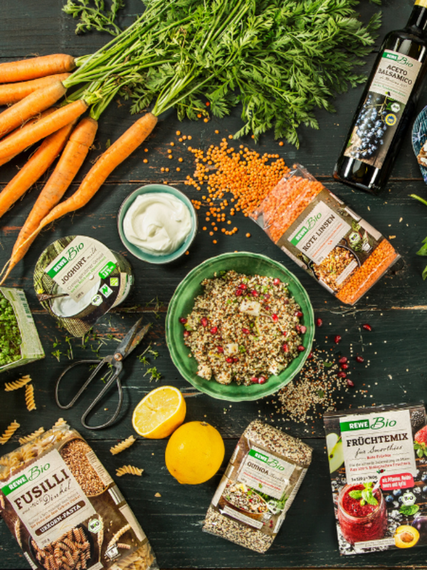 Auf einer Holzplatte liegen viele gesunde REWE-Bio Prokukte. Von Obst und Gemüse über Nudeln, Linsen, Wurst und Hüttenkäse ist alles dabei