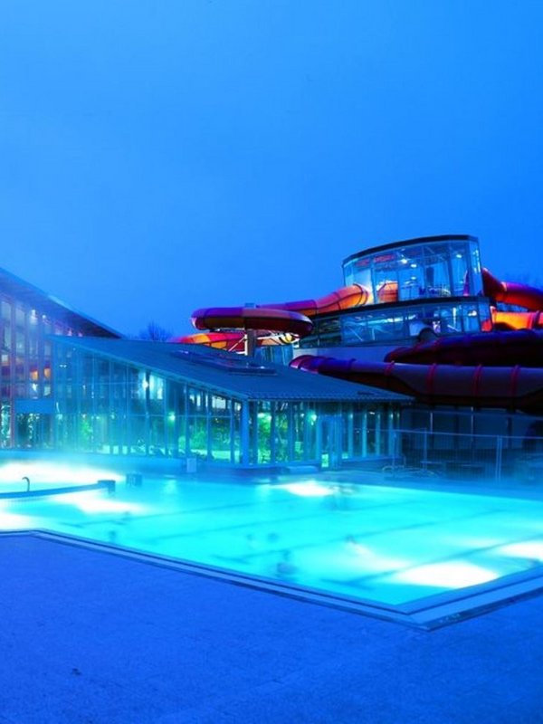 Der Außenpool vom Wonnemar ist im Vordergrund vor dem Gebäude blau beleuchtet. Das Gebäude im Hintergrund hat eine Glasfront und auf der rechten Seite einen Rutschenbereich. Der Himmel ist dunkel, da das Bild abends bei Dämmerung aufgenommen wurde. 