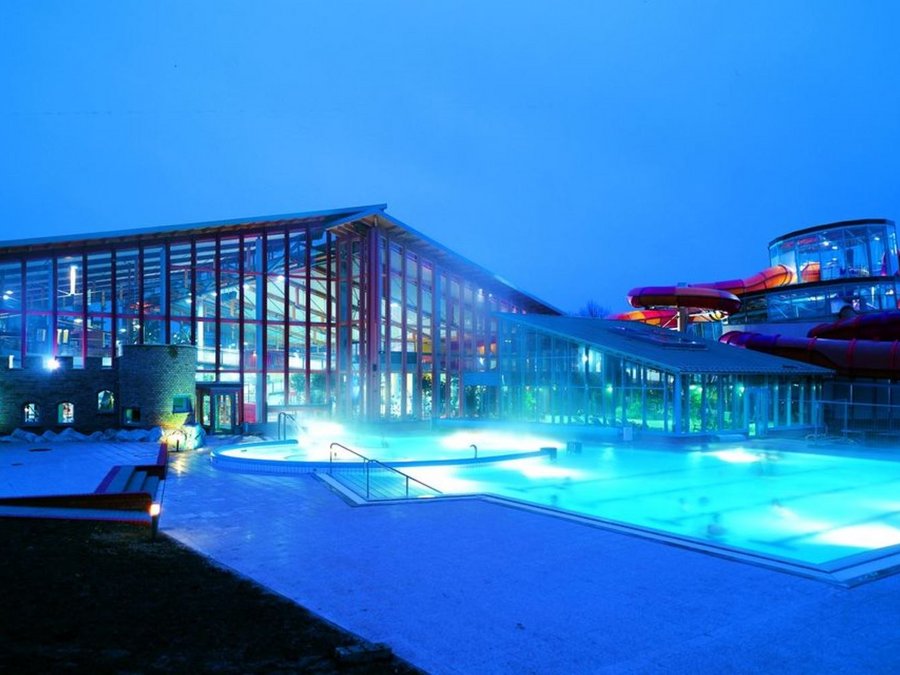 Der Außenpool vom Wonnemar ist im Vordergrund vor dem Gebäude blau beleuchtet. Das Gebäude im Hintergrund hat eine Glasfront und auf der rechten Seite einen Rutschenbereich. Der Himmel ist dunkel, da das Bild abends bei Dämmerung aufgenommen wurde. 