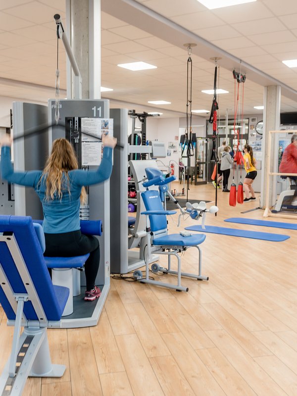 In einem Fitnessraum trainiert eine Frau an einem Kabelzug. Im Hintergrund sind noch weitere Geräte zu sehen. 