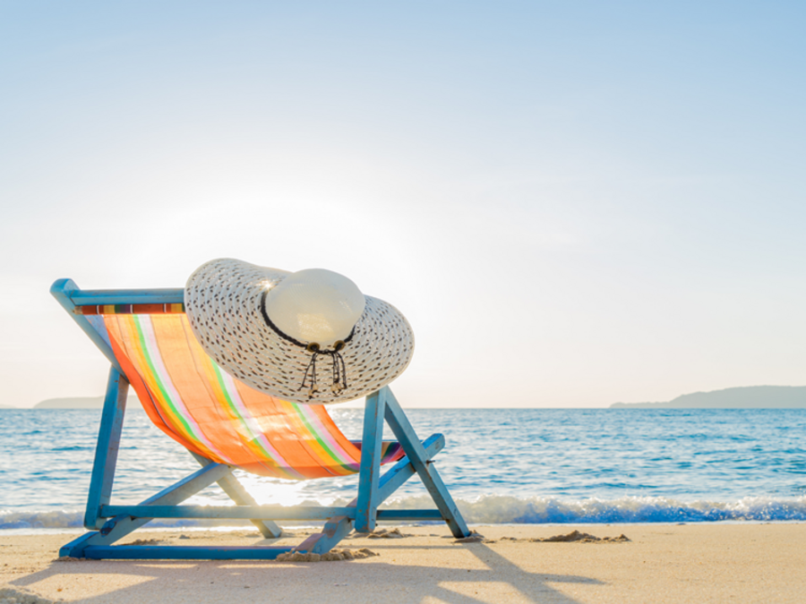 Auf dem Bild steht ein Sonnerstuhl, an dem ein Sonnenhut hängt auf einem weißen Sandstrand,direkt a blauen Meer, die Sonne scheint