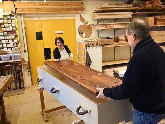 Ein Frau steht auf der einen Seite eines neu gebauten Sarges und ein Herr auf der gegenüberliegenden langen Seite. Sie legen den Deckel auf dem Sarg.