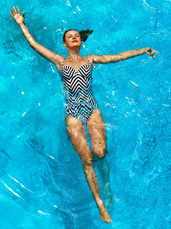 Eine Frau im gemusterten Badeanzug schwimmt auf dem Rücken in blauem Wasser. 