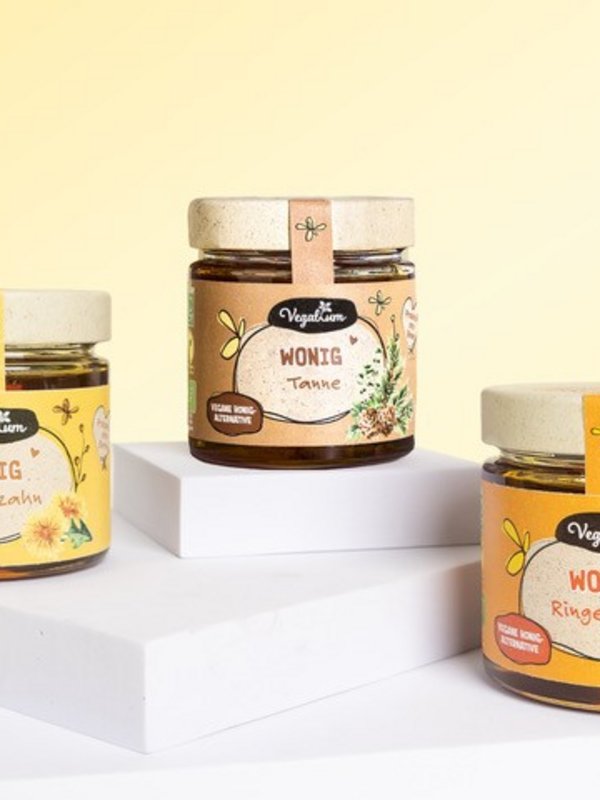 Auf einem weißen Podest sind drei verschiedene Gläser Honig mit gelblicher Banderole und hellem Deckel. 