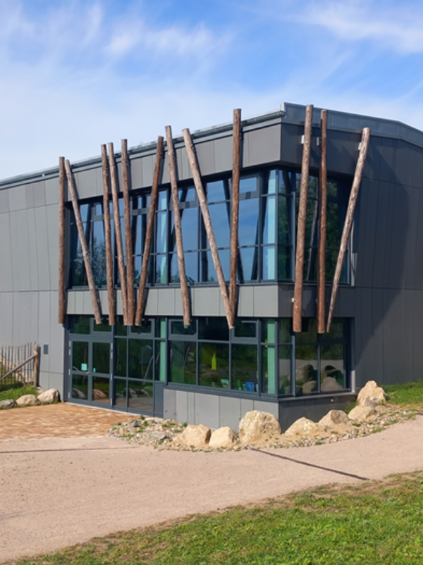 Modernes, zweistöckiges Gebäude mit dunkler Metallfassade und großen Glasflächen. Es hat senkrecht angebrachten Holzstämme an der Front. Das Gebäude steht in einer grünen, naturnahen Umgebung mit Wegen, Grasflächen und einigen Felsen.