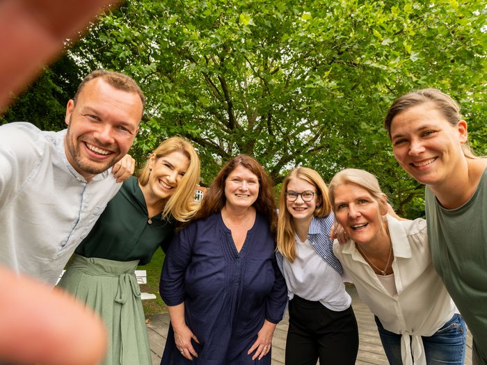 Fröhliche Mitarbeitende in Selfiepose lächeln in die Kamera. Im Hintergrund grüne Bäume.
