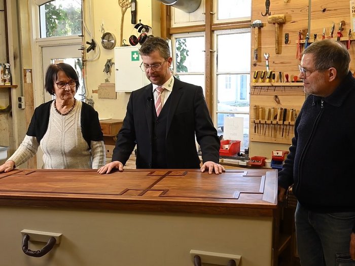 Eine Frau steht an der langen Seite eines geschlossenen Sarges 1 Hand auf dem Sarg, rechts daneben steht ein Herr und hält beide Hände auf den Sarg. An der Stirn des Sarges, auf der rechten Seite steht ein weiterer Herr. Es scheint sich um Mitarbeiter des Unternehmens zu handeln.