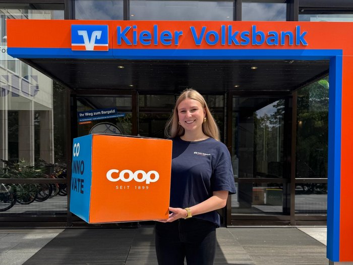 Blonde junge Frau hält einen orange farbenen Würfel mit dem weißen coop Logo in die Kamere. Hinter ihr ist der Eingang in eine Kieler Volksbank, was man an dem Logo und großen Schriftzug über ihr erkennnt.