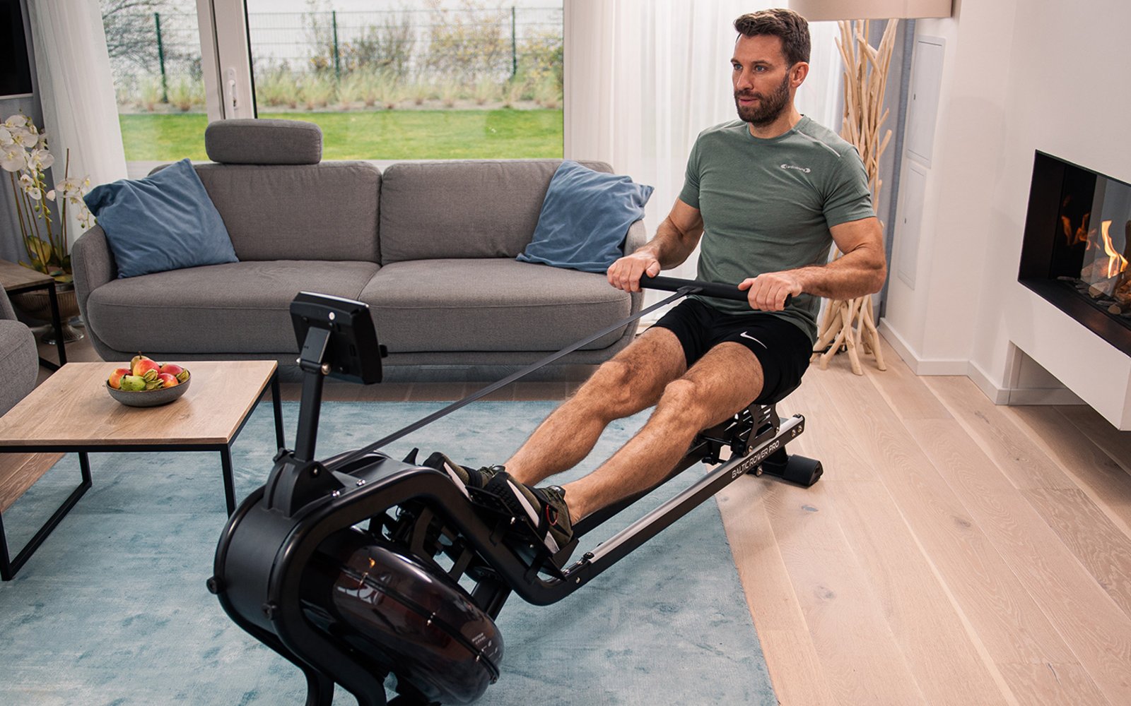 Ein Mann sitzt auf einem Ruder Ergometer in seinem Wohnzimmer. Im Hintergrund ist ein Kamin und ein Sofa. 
