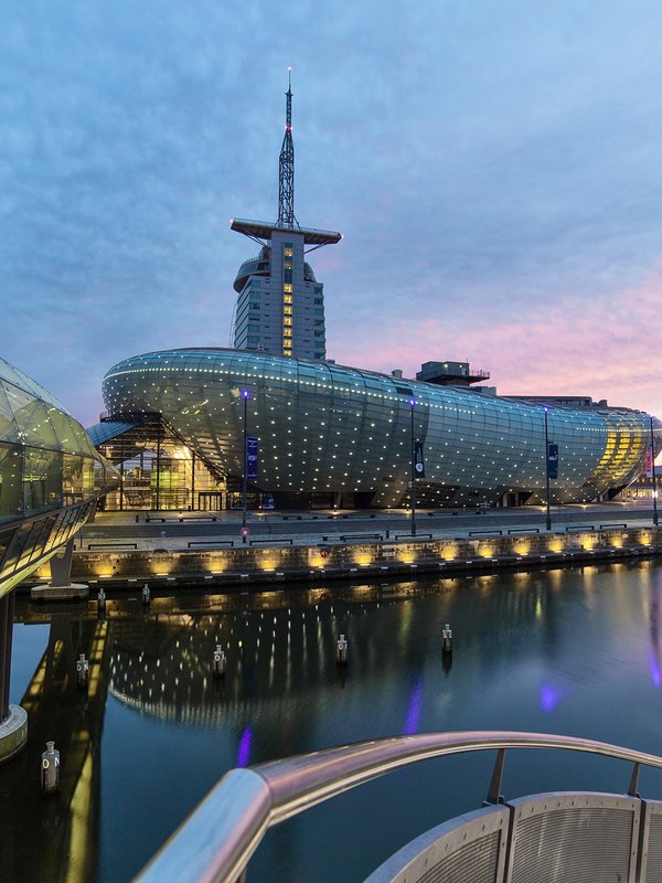 Das Klimahaus Bremerhaven, ein modernes Gebäude mit einer markanten Architektur, das sich durch eine futuristische Fassade und große Fensterflächen auszeichnet. Das Gebäude ist in einer bogenförmigen Struktur gebaut, die an eine wellenartige Bewegung erinnert, und reflektiert die nachhaltige Bauweise und das Thema Klimaschutz. Das Bild wurde abends bei Dämmerung gemacht und entsprechend dunkel ist das Bild. Licht erscheint durch die Fensterfassade. 