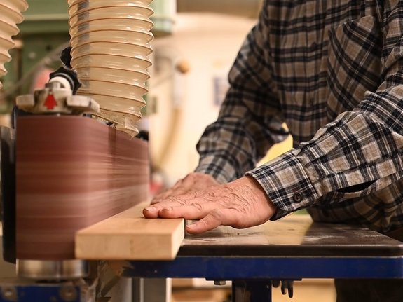 Zwei Hände halten ein Stück Holz an einer Schleifmaschine. Es ist keine Person auf dem Bild zu sehen, nur ein karierter Ärmel.
