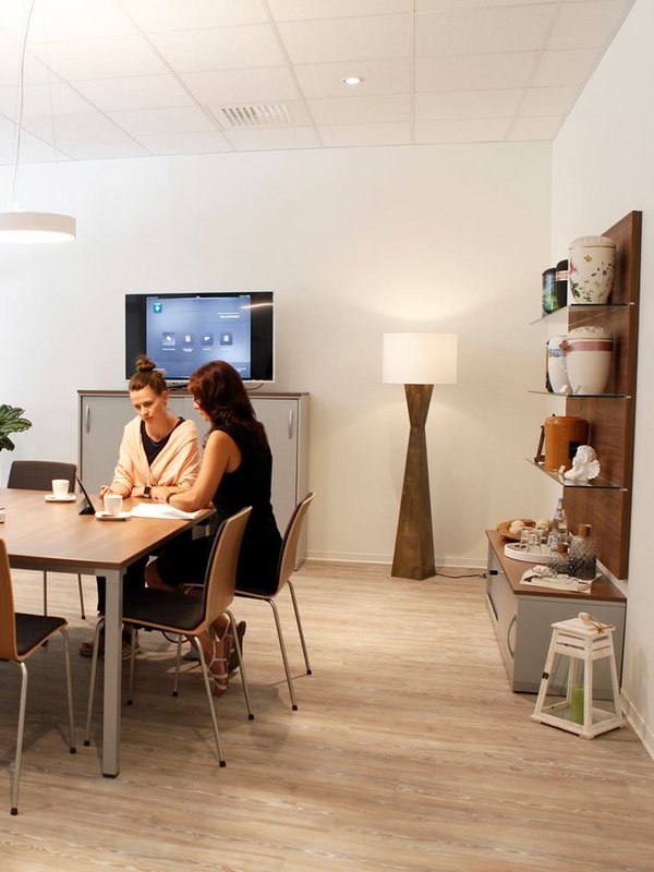 Zwei Frauen sitzen an einem Besprechungstisch in einem modernen, hellen Büro. Sie unterhalten sich und arbeiten gemeinsam an Dokumenten. Im Hintergrund stehen ein Fernseher, eine Stehlampe und Regale mit Geschirr und Dekoration.