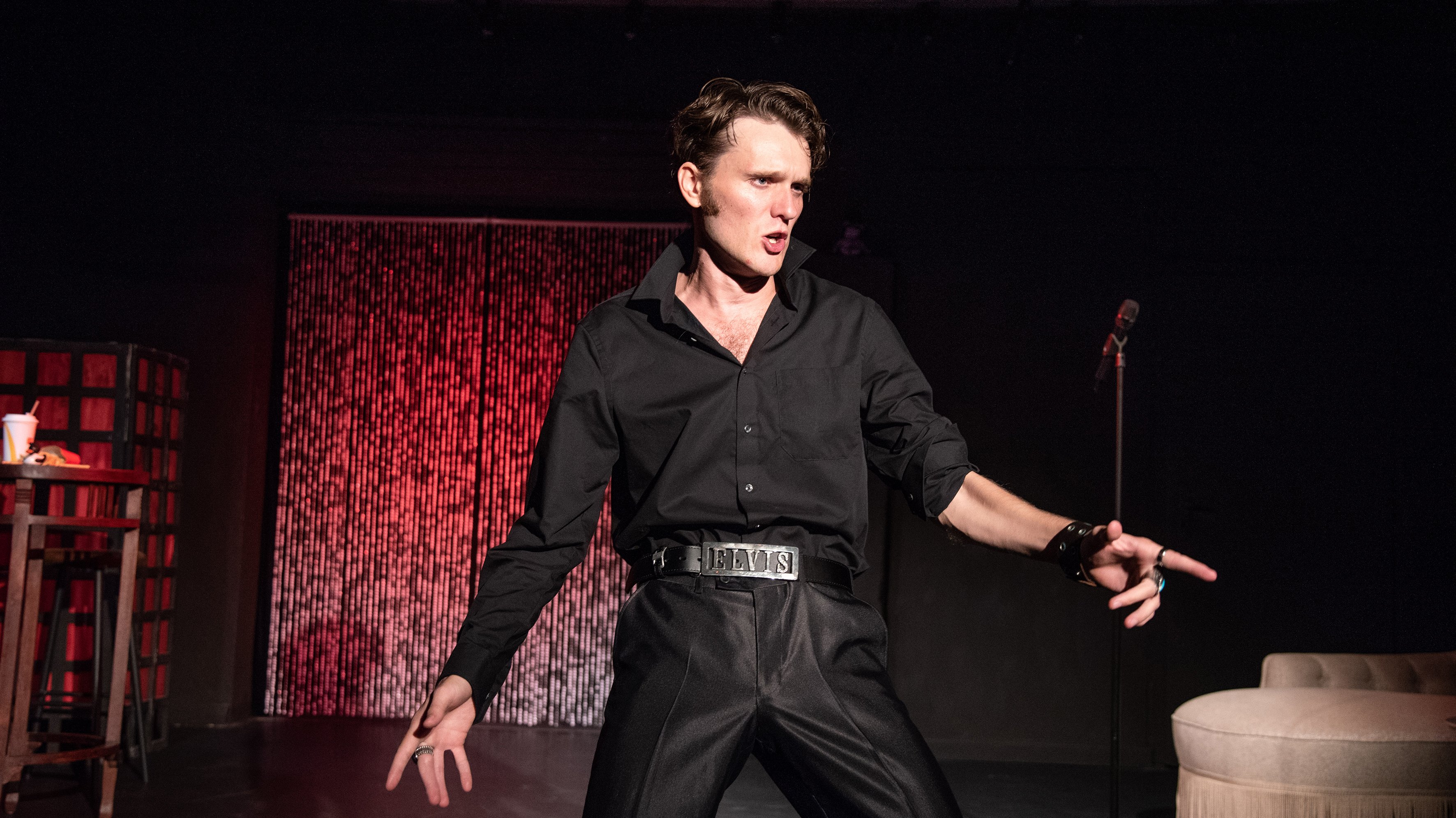 Elvis Darsteller steht in typischer Elvis Pose, Arme etwas ausgebreitet, Finger gespreizt. Er trägt ein schwarzes Hemd und eine schwarze Hose. Im Hintergrund ist ein Lamettavorhang und ein Sofa aus den 60ern