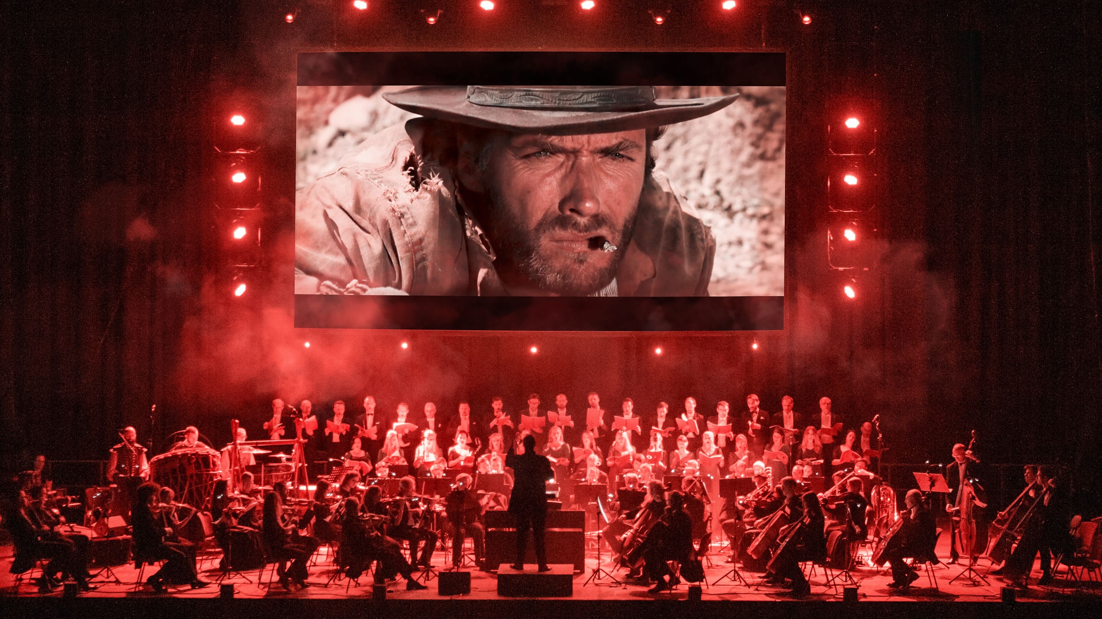 Auf dem Bild ist eine Leinwand mit 3 roten Balken links und rechts und oben. Darunter ist das Gesicht eines Cowboys. Im unteren Bereich ist ein Orchester auf der Bühe, der Dirigent steht in der Mitte