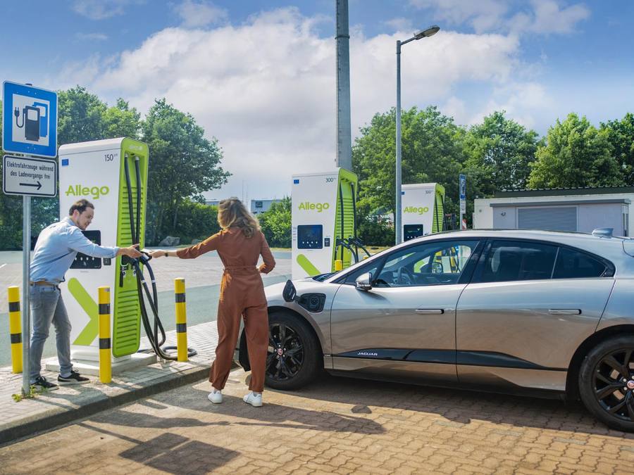Eine Frau und ein Mann bedienen eine Ladesäule von Allego und schließen gerade das Ladekabel vom silbernen Auto an. Im Hintergrund ist blauer Himmel und etwas Wolken zu sehen. 