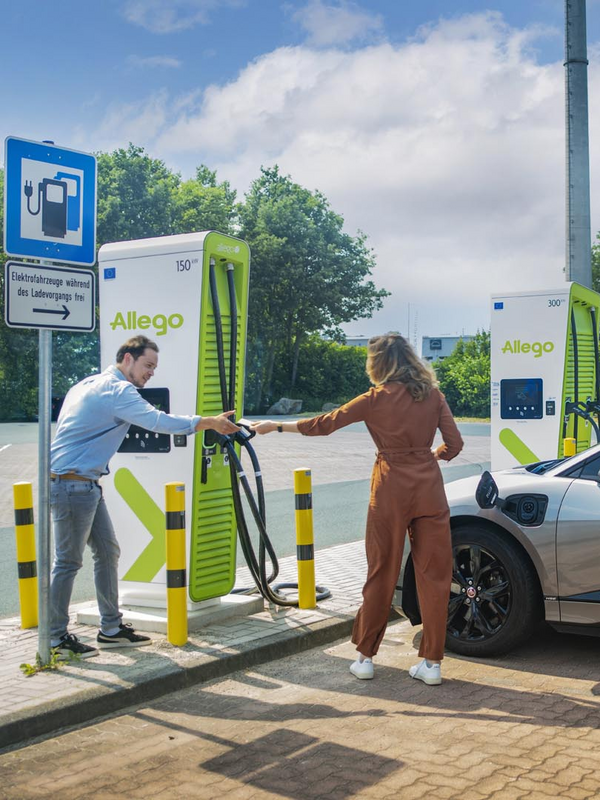 Eine Frau und ein Mann bedienen eine Ladesäule von Allego und schließen gerade das Ladekabel vom silbernen Auto an. Im Hintergrund ist blauer Himmel und etwas Wolken zu sehen. 