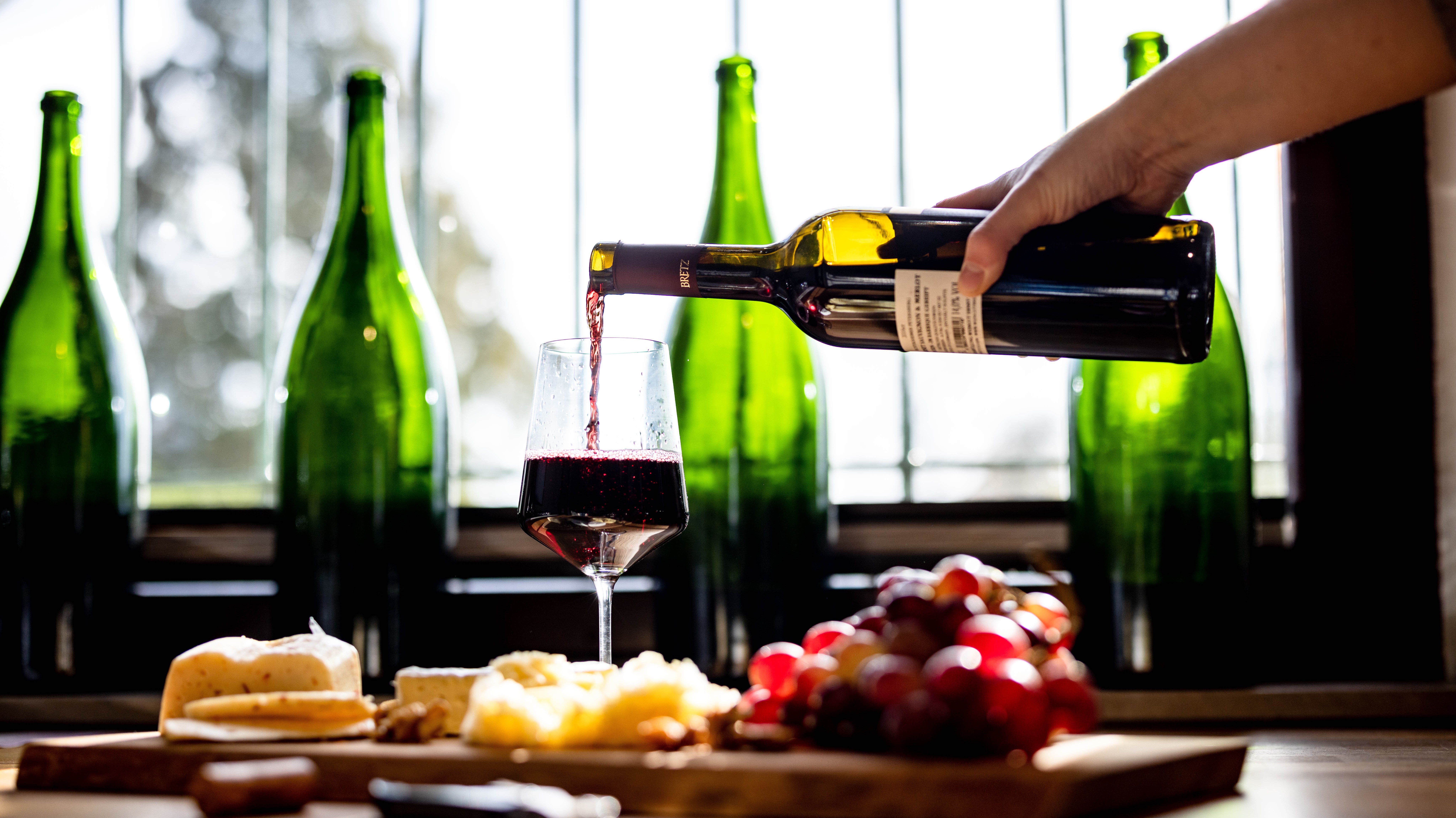 4 grüne Flaschen Wein im Hintergrund. Im Vordergrund ein Weinglas zur Hälfte mit Rotwein gefüllt. Eine Hand schenkt derweil noch mit einer Flasche in das Glas ein. Das Glas steht auf einem Holzbrett, welches mit Brot, Weintrauben und Käse bestückt ist