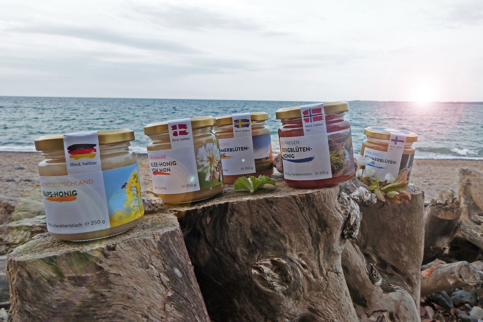 Drei Gläser mit Honig stehen auf einem Baumstamm. Im Hintergrund ist Meer und Sonne. 