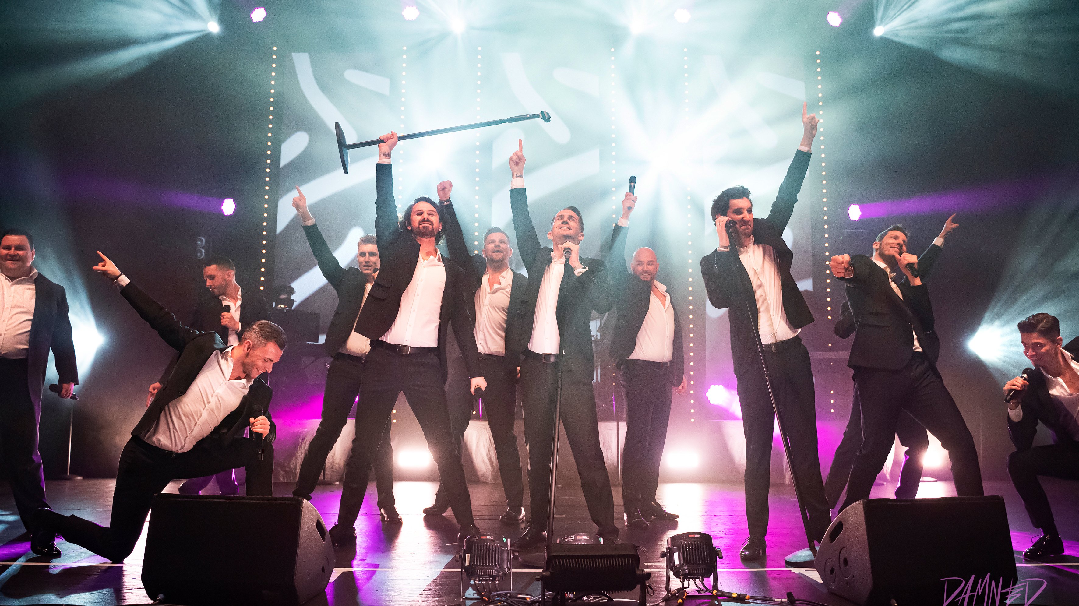 12 Tenöre stehen auf der Bühne und heben jeweils einen Arm in die Luft, darüber ist helles Scheinwerferlicht