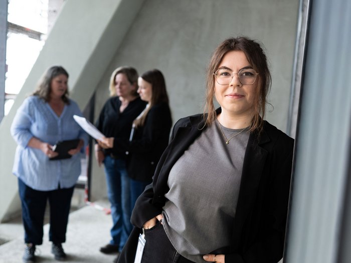 Junge dunkelhaarige Frau lehnt an einem Stahlträger und schaut in die Kamera. Im Hintergrund Rohbauatmosphäre und drei Frauen stehen im Halbkreis und diskutieren über ein Dokument in der Hand.