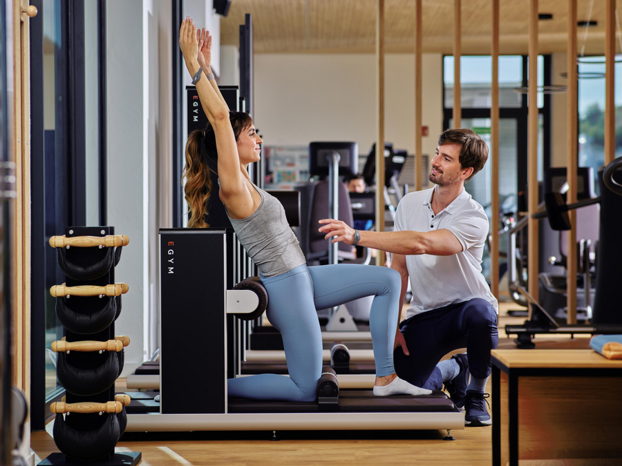 Eine Frau trainiert an einem Trainingsgerät und wird von einem Trainer, der davor kniet, angeleitet. 