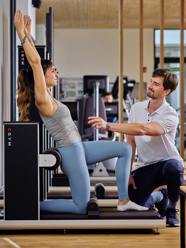 Eine Frau trainiert an einem Trainingsgerät und wird von einem Trainer, der davor kniet, angeleitet. 