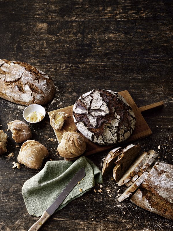 Brot und Brötchen liegen auf einer großen Holzplatte. 2 Brote sind noch als Laib und 2 Brote bereits geschnitten zu sehen. Auch die Brötchen sind teilweise in der Mitte gebrochen. Der Hintergrund wirkt sehr dunkel.