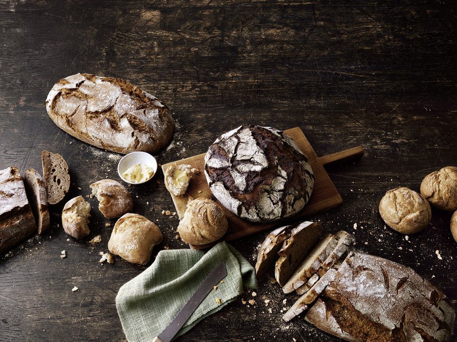 Brot und Brötchen liegen auf einer großen Holzplatte. 2 Brote sind noch als Laib und 2 Brote bereits geschnitten zu sehen. Auch die Brötchen sind teilweise in der Mitte gebrochen. Der Hintergrund wirkt sehr dunkel.