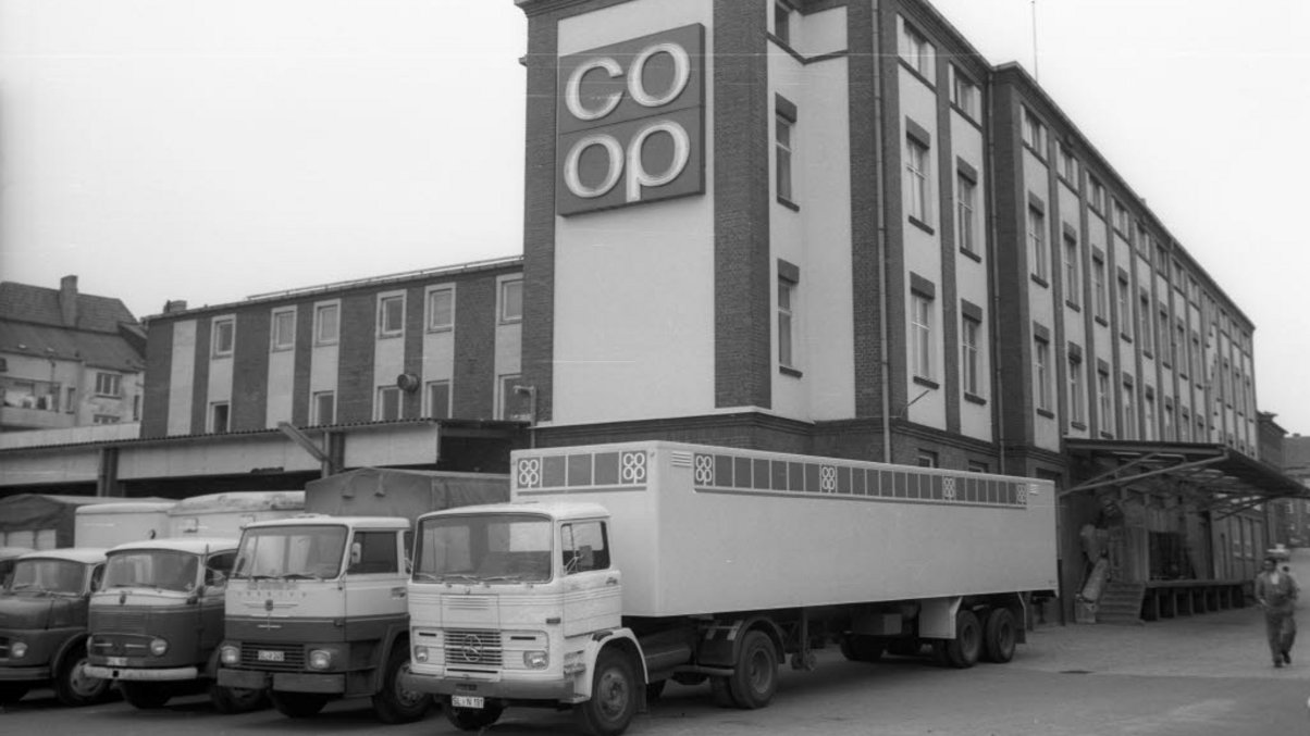Schwarz-Weiß-Aufnahme der früheren coop Zentrale mit großem coop Logo am Eckturm des Gebäudes, or dem eine Reihe LKWs stehen