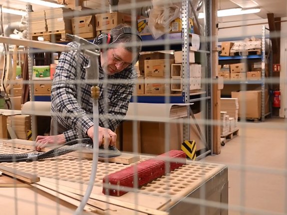 Ein Mann schleift in einer Werkstatt mit Geräuschschutz auf den Ohren ein langes Stück Holz