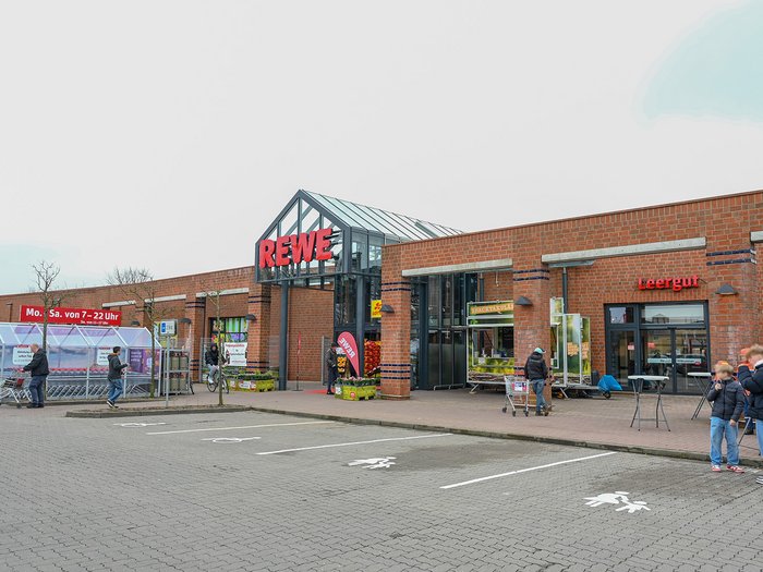 Die Außenfront des Supermarktes aus Klinker trägt das große REWE-Logo. Menschen sind im Vordergrund zu sehen.