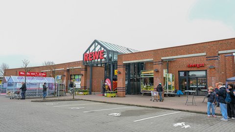 Die Außenfront des Supermarktes aus Klinker trägt das große REWE-Logo. Menschen sind im Vordergrund zu sehen.