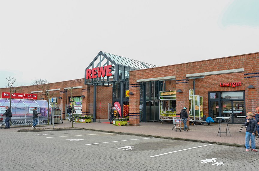 Die Außenfront des Supermarktes aus Klinker trägt das große REWE-Logo. Menschen sind im Vordergrund zu sehen.