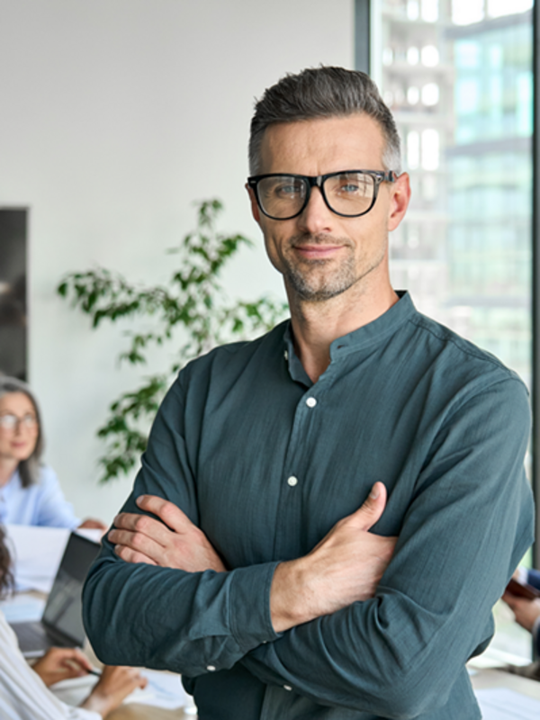 Ein Mann mit Brille steht im Mittelpunkt des Bildes mit verschränkten Armen. Im Hintergrund ist ein Besprechungsraum in dem vier weitere Menschen sitzen. 