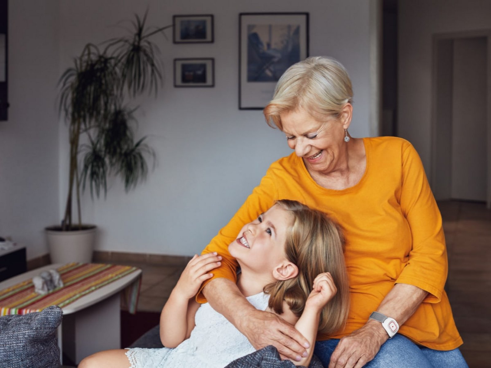 Ein Kind liegt lachend im Arm der Oma. Sie sitzen auf einer Couch. Im Hintergrund ist das Wohnzimmer mit Bildern und einer Pflanze. 