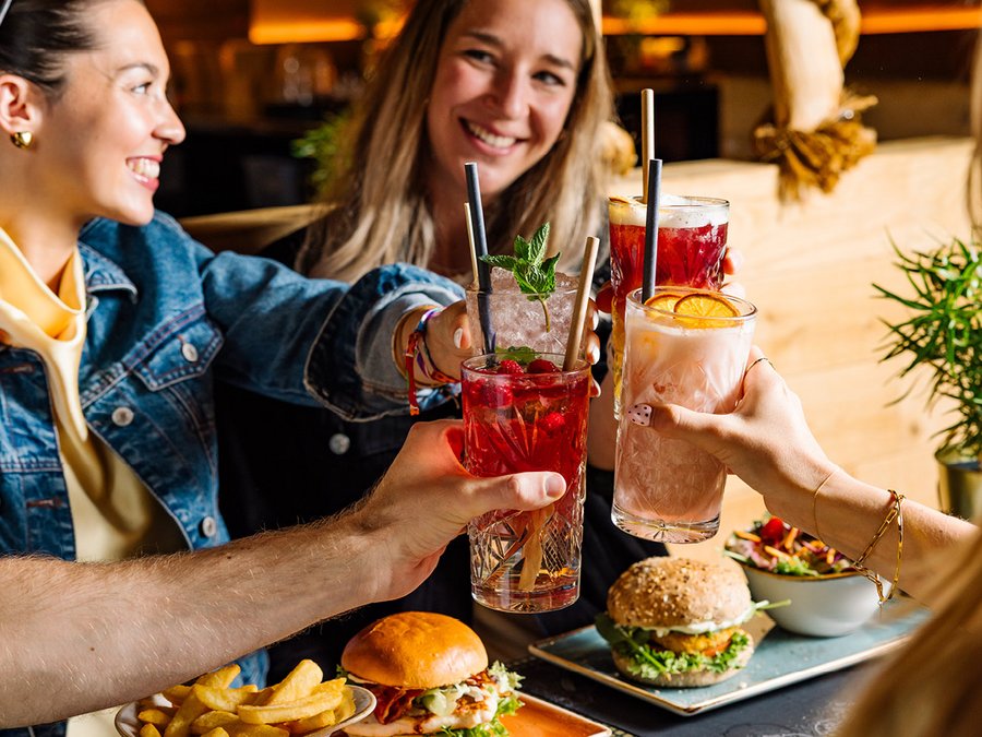 2 Frauen stoßen mit je 1 Glas mit 2 weiteren Personen, von denen nur die Hände mit je 1 Glas zu sehen ist an. Sie lächeln. Pommes und Burger liegen auf einem Teller vor den 4 Personen.